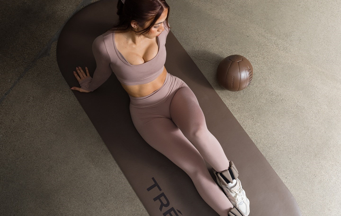 Woman in mauve workout set sitting on an exercise mat with ankle weights and a medicine ball nearby.