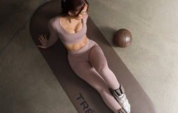 Woman in mauve workout set sitting on an exercise mat with ankle weights and a medicine ball nearby.