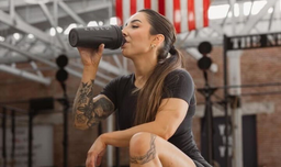Athletic woman kneeling in a gym, drinking from a Kaged shaker bottle during a workout session.