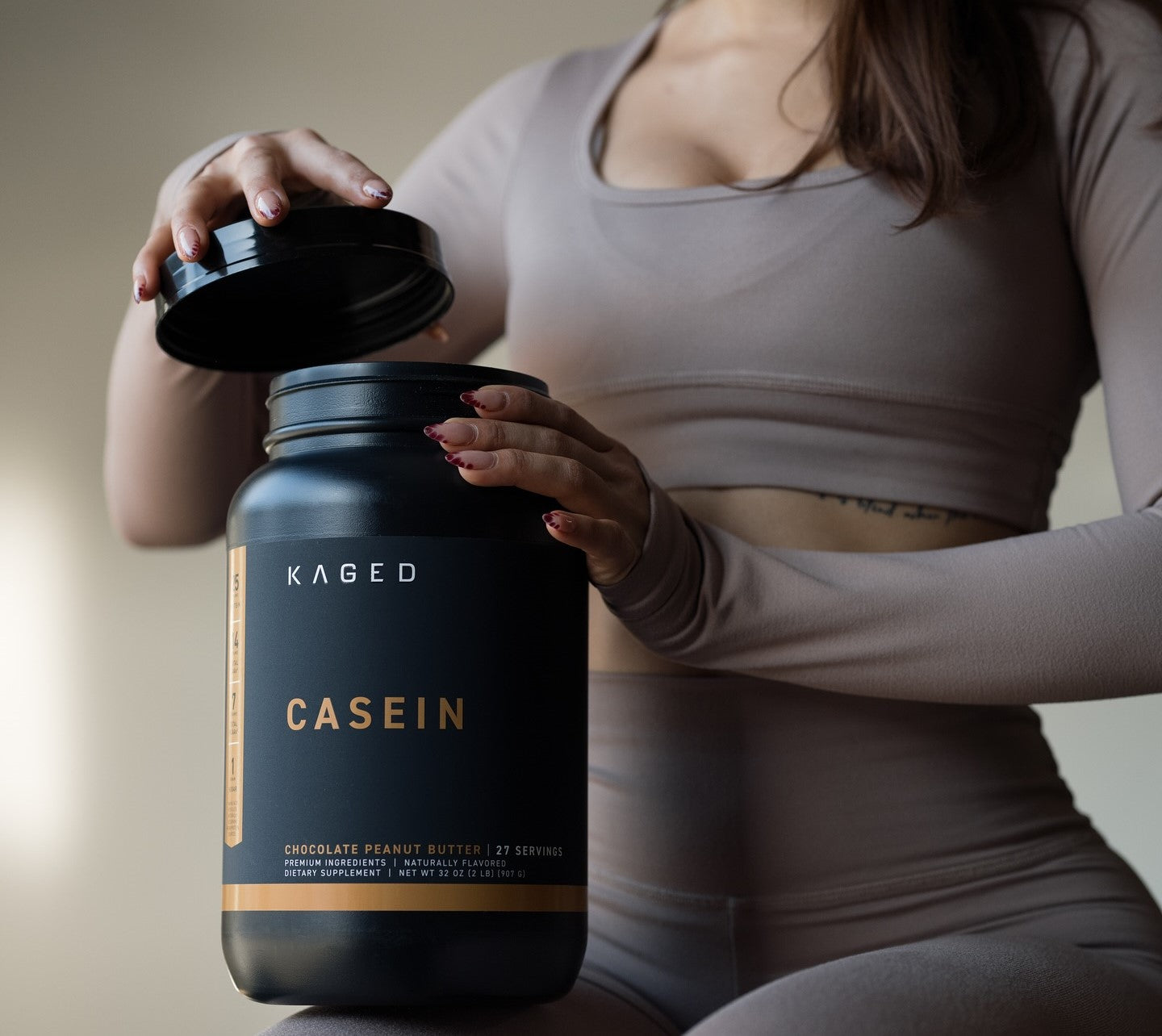 A woman in active wear, opening a Kaged Casein protein powder container.