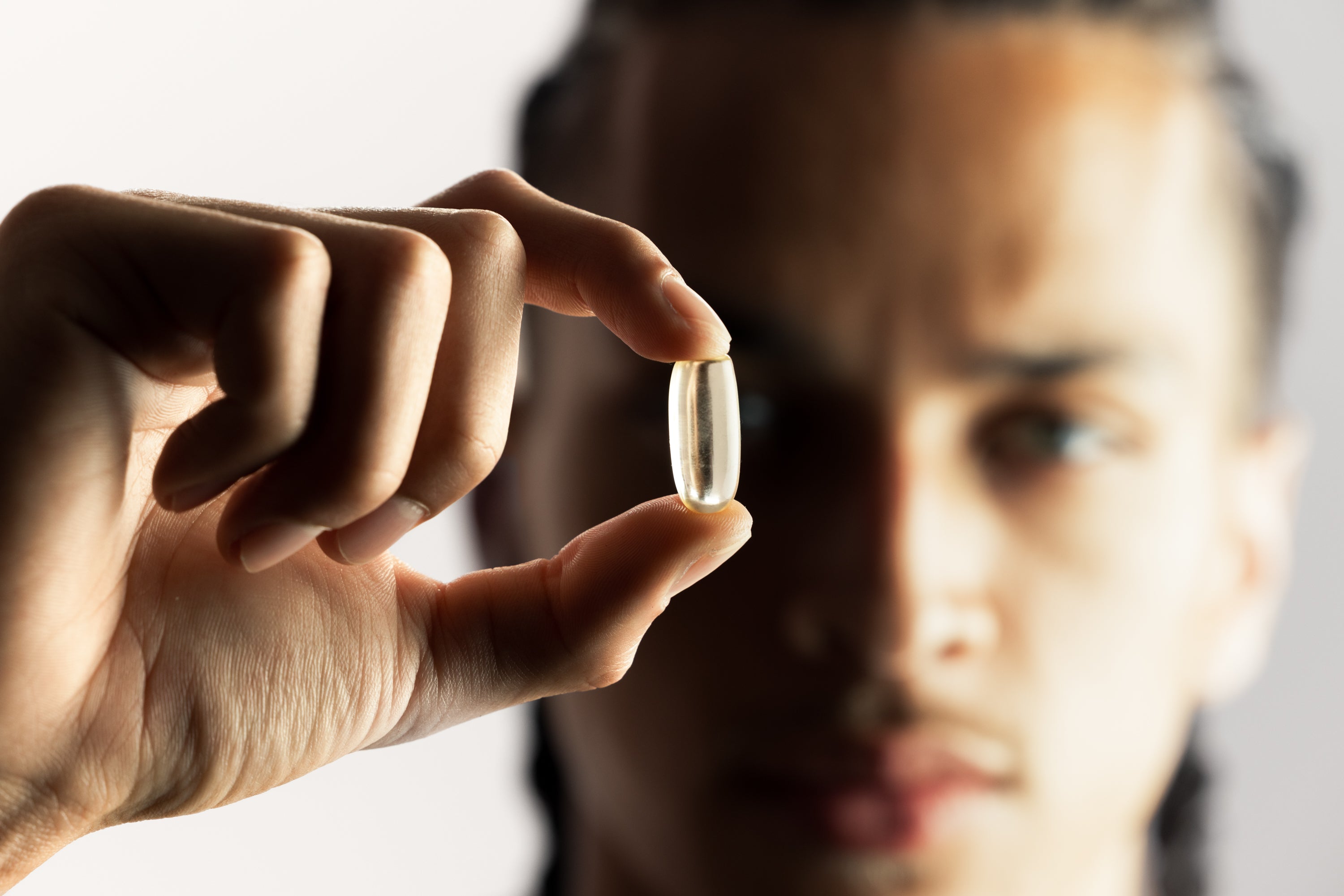 Person holding a clear supplement softgel between fingers in front of a blurred face to emphasize transparency