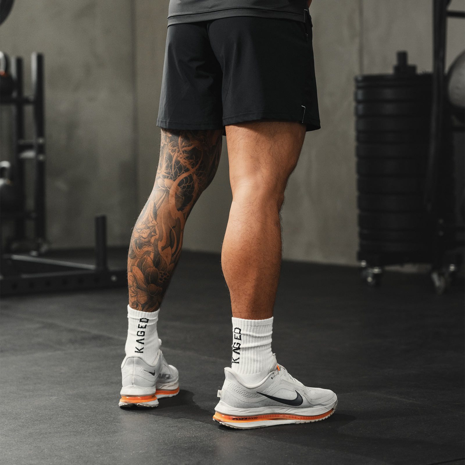 Rear view of Ascent Performance Socks in white on a model, showing ribbed crew height and KAGED branding on the cuff in a gym