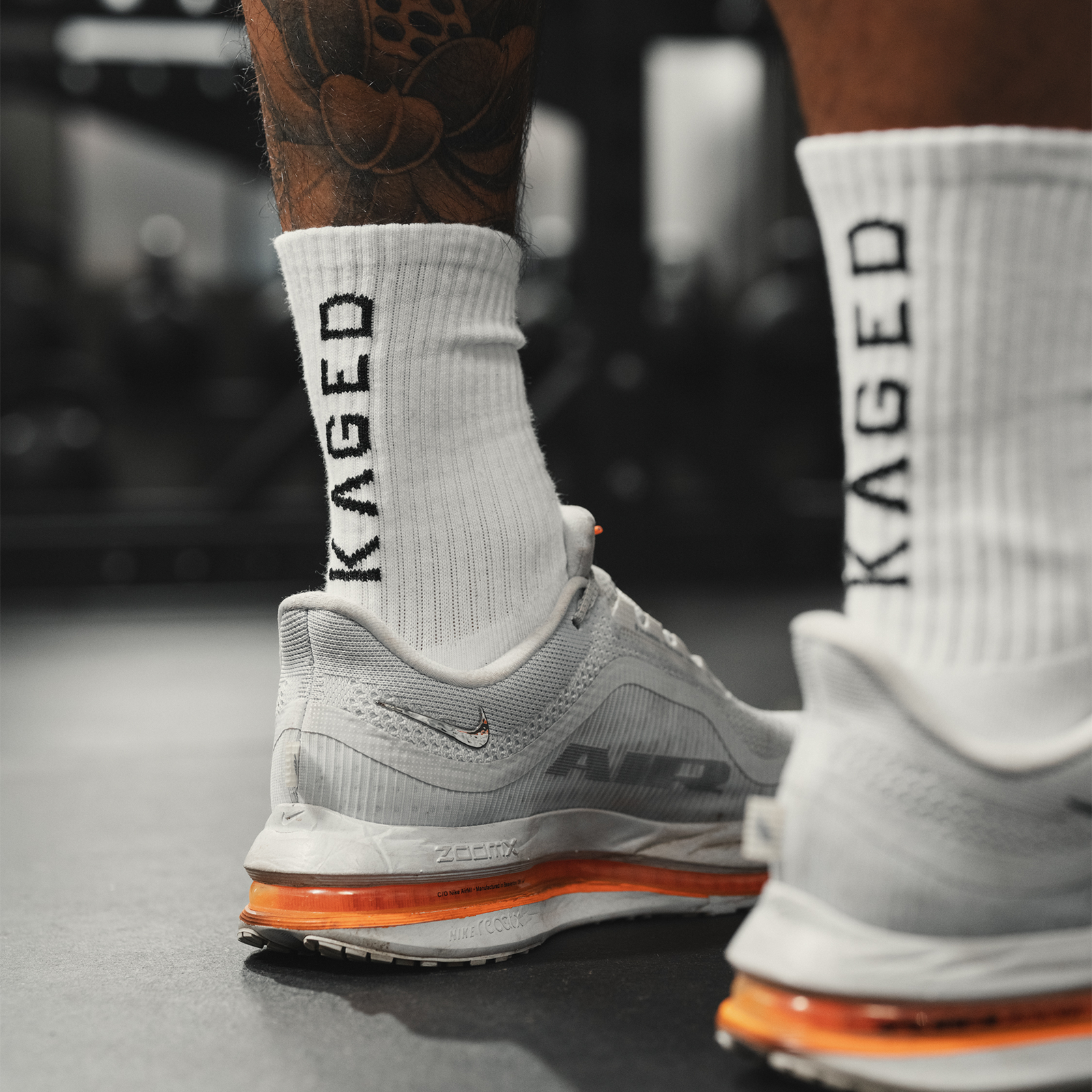 Rear three-quarter view of Ascent Performance sock in white showing KAGED logo, ribbed crew and reinforced heel in gym