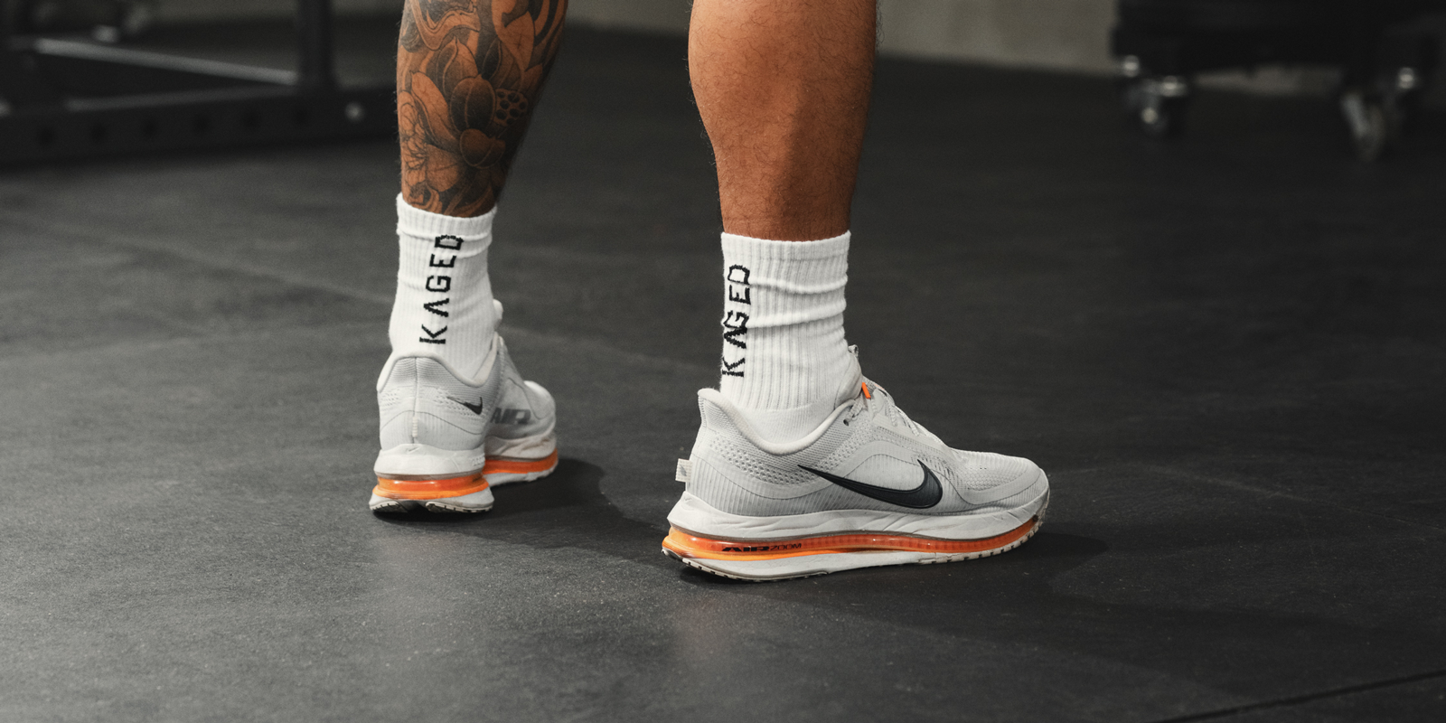 Rear three-quarter view of white Ascent Performance crew socks on model showing KAGED logo and ribbed cushioning on a gym floor
